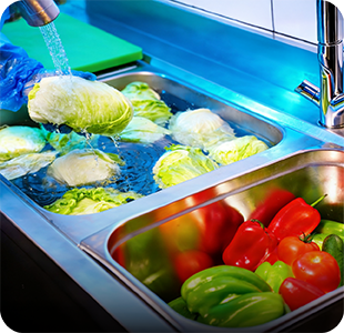 Lettuce being washed under running water beside trays of fresh peppers